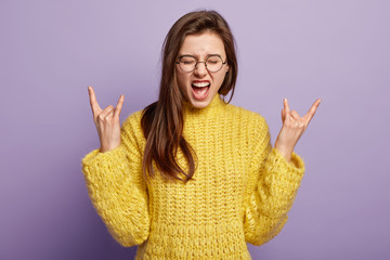 Emotive European woman makes horns with fingers, raises hands, exclaims loudly, wears yellow jumper, poses over purple background. Impressed lady shows rock n roll gesture. I like heavy metal