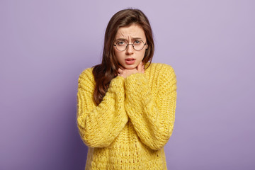 Isolated shot of displeased young female keeps both hands on neck, suffers from cough, has asthmatic fit, wears round glasses and yellow sweater, models over purple wall. Thore throat, illness