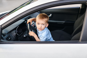 A blond boy with blue eyes sits in the front seat behind the wheel of a white car.