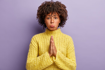 Unhappy woman pleads for forgiveness, keeps palms together, has regretful facial expression,...