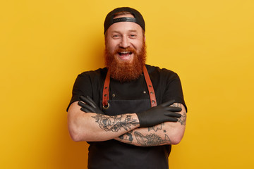 Cheerful male chef in black apron, t shirt and gloves, has thick long ginger beard, keeps arms folded, laughs sincerely, has break after cooking, talks with colleagues in restaurant, shares new recipe - obrazy, fototapety, plakaty