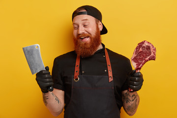 Shot of pleased male meat chopper glad to have sharp cleaver for cutting meat, dressed in black outfit, works at butchers shop or market, has ginger thick beard, isolated over yellow background