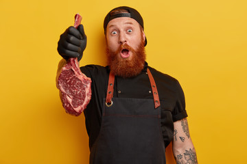 Surprised cheerful male butcher holds fresh raw piece of meat with bone, has red thick beard,...