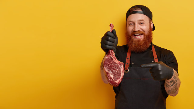 Indoor shot of man works at meat packing plant, points at ribeye steak, involved in butchering, suggests to bake pork for dinner, has ginger beard, models over yellow wall with empty space aside
