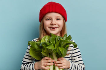 Headshot of smiling little female preschooler holds bok choy greenery, wants to eat fresh spring salad, wears red headgear and striped clothes, being eco friendly, isolated over blue background