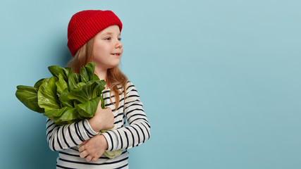 Cute small child in red headgear and striped jumper, carries green salad, looks away, wants to make...
