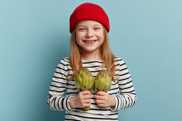 Isolated shot of cheerful lovely girl carries artichoke, helps mother with preparing fresh spring...