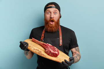 Shocked bewildered bearded man chef with thick beard, holds piece of raw meat on wooden cutting...