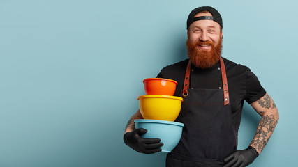 Image of smiling pleased chef holds hand on waist, carries utensils, wears apron and t shirt, being in high spirit for cooking something delicious, stands against blue wall with free space aside