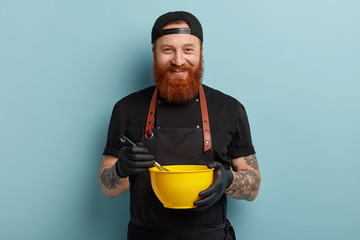 Baking and cooking concept. Happy ginger male chef holds bowl and whipping utensils, whisks eggs in...