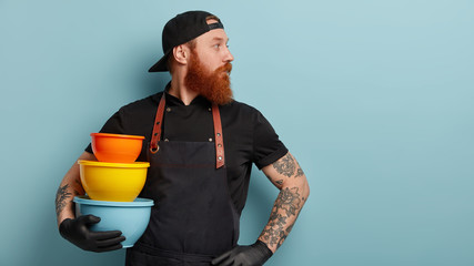 Indoor shot of serious man with thick foxy beard, focused aside with thoughtful expression, carries colorful food containers, poses indoor over blue background, works in restaurant at kitchen