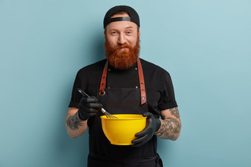 Self confident unshaven guy whisks eggs in bowl, bakes delicious cuisine, wears black clothes, has...