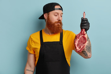 Photo of serious red haired bearded male chef holds piece of meat, going to prepare pork on grill,...