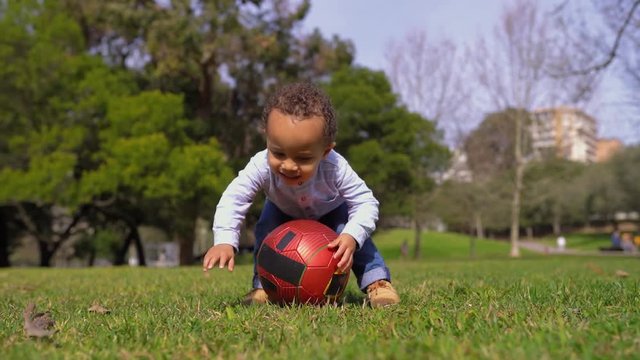 Little mixed-race boy in blue shirt nd jeans playing with red ball on grass in park. Low camera position. Family, game concept