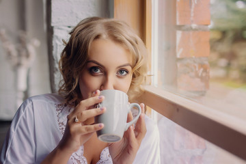 Beautiful blonde woman standing next to a window, wearing night clothes and drinking coffee