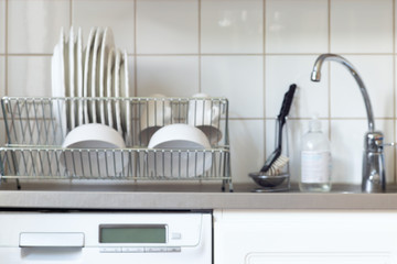 Blur kitchen background with dishwashing machine, water tap, sink, dish rack and plates cups on it,...