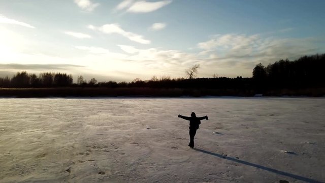 Aerial view: One man is skating on the ice of frozen river during beautiful sunset. Drone is following him and making the video.