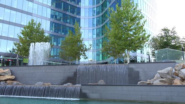 A tall, windowed office building in an urban area on a sunny day - a small artificial waterfall in the foreground