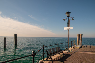 Scenic view of a dock on the shore of Lake Garda with a bench and cloudy blue sky, Lazise, Veneto, Italy