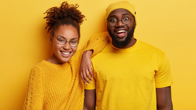 Photo of cheerful dark skinned young lady and man stand closely, enjoy spare time, listens funny news from interlocutor, wear spectacles and yellow clothes. People, emotions, ethnicity concept