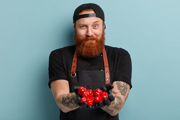 Strict skilled ginger chef has cheerful expression, raises eyebrow, wears black cap and apron,...