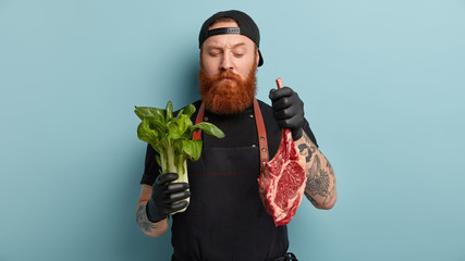 Indoor shot of male cook holds green bok choy in one hand and raw meat in other, going to bake...