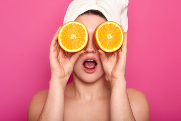 Studio shot of pleasant looking young shocked European woman covers eye with oranges, has white towel on head. Model with clear skin poses in studio isolated over pink background. Beauty concept.