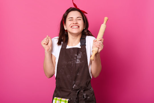 Baker woman holds baking rolling pin, wears brown apron, white t shirt. Young girl bends elbows and shouts yes with closed eyes, female maskes cake first time. Cook isolated on pink background.