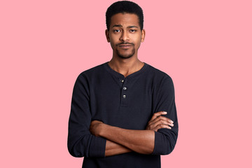 Young dark skinned man with calm expression, short hairstyle and beard, dresses casual shirt and...