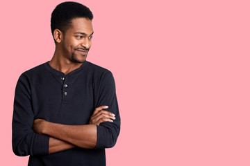 Portrait of confident charismatic young handsome African American male in black casual shirt with hands crossed on chest. Guy looks smiling aside. Attractive man with short hairstyle and beard poses.