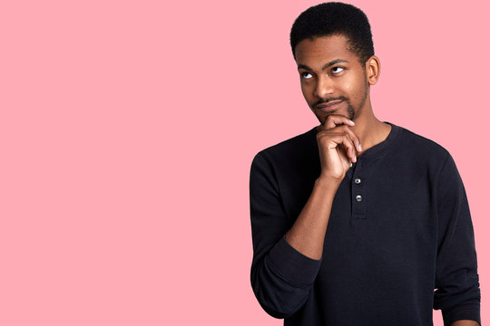 Closeup portrait of attractive thoughtful African guy. Young handsome man looks aside, keeps hand on chin and thinks about his dream, isolated on rose background. Copy space for your advertisment.