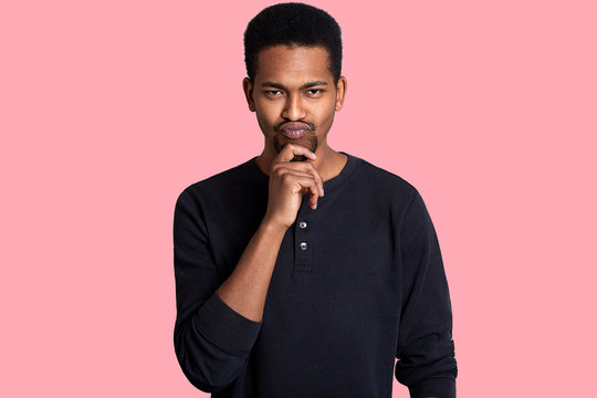Single handsome fit black man, keeps hand on chin. Guy has thoughtful fecial expressions, has interesting idea and thinks how to bring it to life. Handsome male isolated over pink background.