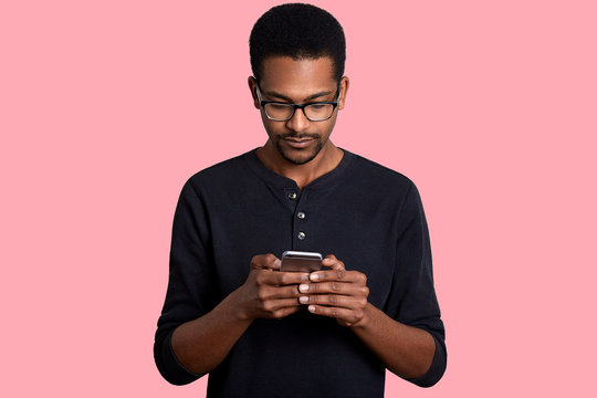 Confident Young African Man Looks At Smart Phone And Sends Text Messege, Connected To Wireless Internet, Stands Against Pink Background. Dark Skinned Guy Wears Black Shirt And Glasses Poses In Studio.