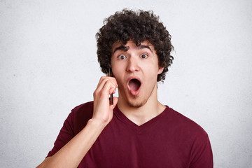 Young dark haired handsome man speaks on phone isolated over white background, has surprised expression. Attractive guy with curly hair stands with opened mouth, hears shocking news from friend.