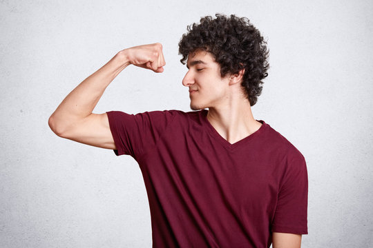 Positive dark haired teenager, dressed casually, shows biceps with satisfied facial expressions, demonstrates how cool he is. Student with curly hair looks at his muscules. Sport and healthy concept.