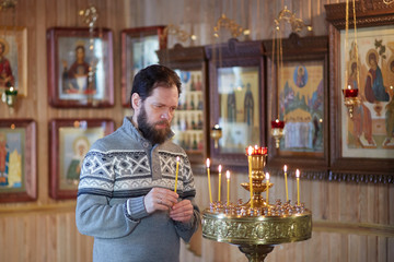 A Russian man with a beard stands in an Orthodox Church, lights a candle and prays in front of the icon.
