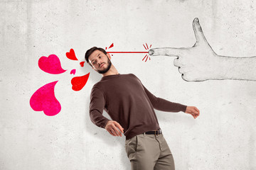 Obraz na płótnie Canvas Young handsome man in casual clothes standing in half-turn near wall and leaning back as if from hand-drawn finger gun shooting pink hearts at him.