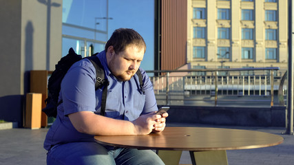 Fat young man scrolling smartphone, sitting in outdoor cafe, lazy lifestyle