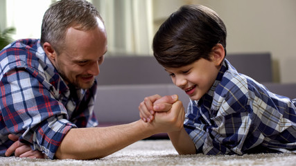 Cheerful father and son arm-wrestling on floor, having fun together, rival