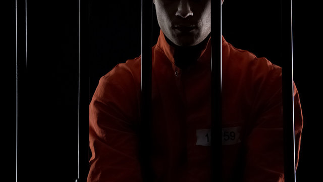 Prisoner in orange uniform standing behind bars, punishment for committed crime