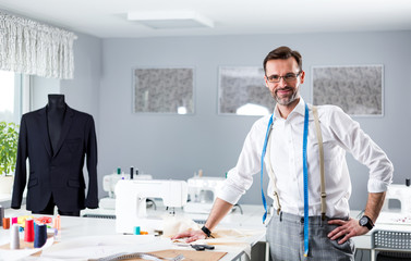 Smiling tailor shop owner standing above workshop desk, fashion design business