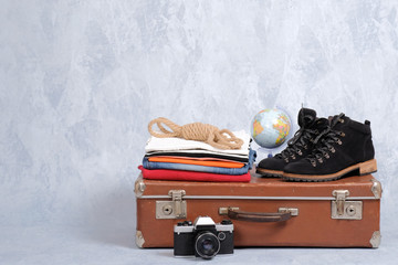 Retro leather suitcase with travel accessories on grey background. Stack of clothes, film photo camera, suede shoes, globe. Travel concept, banner mockup with copy space