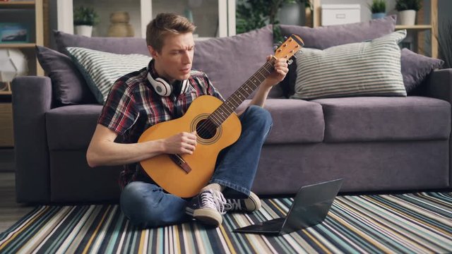 Student is learning to play the guitar watching online lesson in Internet using laptop sitting on floor in living room and playing the musical instrument.