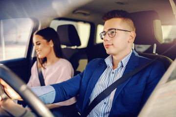 Sophisticated young happy Caucasian couple driving in car. Man holding hands on steering wheel.