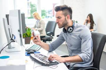Side view portrait of handsome young man with headphones around his neck reading something on smart phone while working on computer in office.