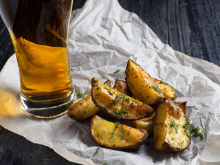 A glass of light beer with a snack in the form of rustic potatoes with dill on a dark wooden background