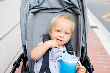 Baby drinking water from special cup sitting in stroller outdoors