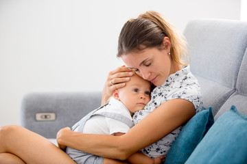 Young mother, sitting at home in sunny living room, cuddling with her toddler baby boy, breastfeeding him