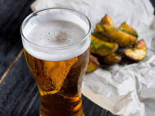 A glass of light beer with a snack in the form of rustic potatoes with dill on a dark wooden background