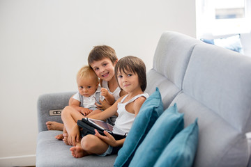 Naklejka na ściany i meble Sweet children, sitting on couch in sunny living room, playing on tablet